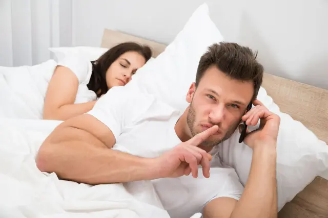 Mężczyzna w łóżku rozmawia przez telefon, robiąc gest ciszy. Obok śpi kobieta. Scena sugeruje zdrada emocjonalna.